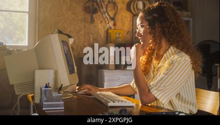 Hispanic Female Software Engineer Programming On Old Desktop Computer In Retro Garage With Random Appliances. Young Woman Starting Fintech Startup Company In Nineties. Nostalgia Concept Stock Photo