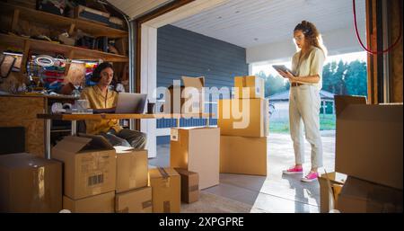Caucasian Male Small Business Owner Using Laptop Computer In Home Garage To Fill Online Orders And Talking To Hispanic Female Manager. Multiethnic Businesswoman Using Tablet To Check Inventory. Stock Photo