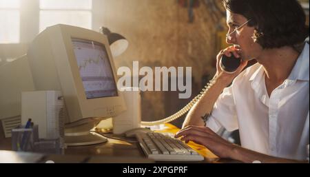 Caucasian Male Trader Talking On Landline Phone And Using Old Desktop Computer In Retro Garage. Young Man Pitching New Promising Stock To Investors. Starting Investment Management Fund in Nineties. Stock Photo