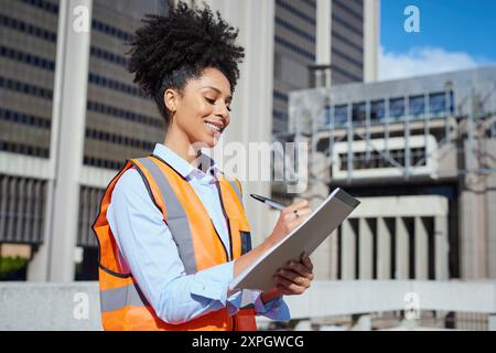 Female Construction Worker in Safety Vest Smiling and Taking Notes Outdoors Stock Photo
