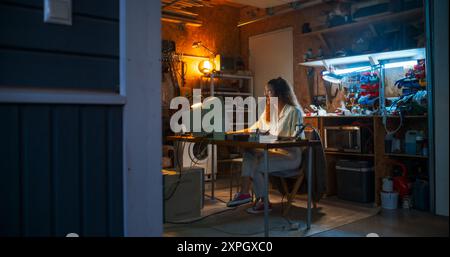 Wide Shot Of Hispanic Female Engineer Using Old Computer In Nineties Retro Garage In The Evening. Intelligent Woman Programming, Working On Innovative Portable Device, Starting Technological Company. Stock Photo