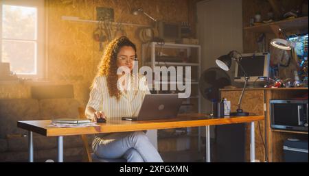 Hispanic Female Programmer Writing Code On Laptop Computer In Garage At Home. Woman Working On Innovative Software As A Service Startup For Business. Digital Entrepreneur Starting Company Concept. Stock Photo