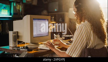 Side View Of Hispanic Female Engineering Student Writing Academic Thesis On Old Desktop Computer In Retro Garage. Future Engineer Focused On Innovative Project For University Of Technology In Nineties Stock Photo