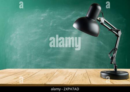 Black desk lamp on wooden table against green school chalkboard background. Back to school concept with copy space. Empty background. Stock Photo