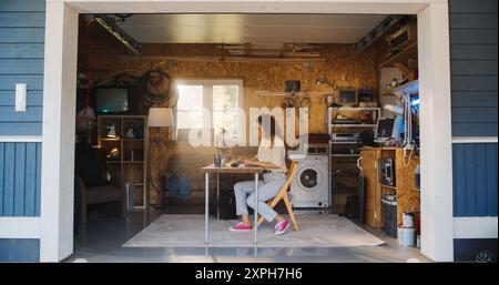 Hispanic Female Software Developer Programming On Laptop Computer In Garage At Home. Focused Young Woman Working On Innovative Online Service Startup Company, Coding. Digital Entrepreneur Concept. Stock Photo