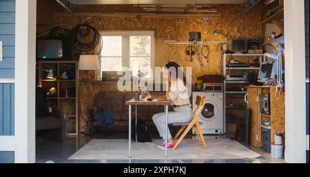 Hispanic Female Software Engineer Programming On Laptop Computer In Garage At Home. Focused Young Woman Working On Innovative Online Service Startup Company, Coding. Digital Entrepreneur Concept. Stock Photo