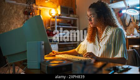 Hispanic Female Engineer Using Old Desktop Computer In Nineties Retro Garage In The Evening. Intelligent Woman Programming, Working On Innovative Portable Device, Starting Tech Startup Company. Stock Photo