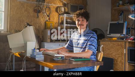 Caucasian Male Software Engineer Programming On Old Desktop Computer In Retro Garage, Looking At Camera And Smiling. Man Starting an Innovative Startup Company In Nineties. Nostalgia Concept. Stock Photo