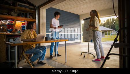 Team Of Young Entrepreneurs Having A Meeting In Home Garage. Hispanic Female Manager Using Whiteboard To Outline Business Plan For Computer Production. Two Male Colleagues Using Laptop And Tablet. Stock Photo