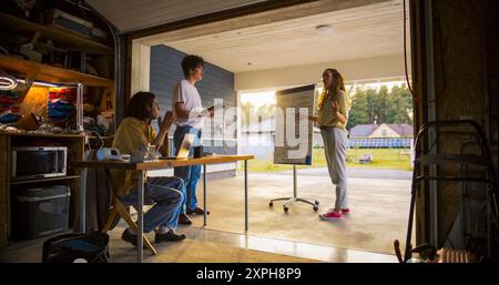 Team Of Young Entrepreneurs Having A Meeting In Home Garage. Hispanic Female Manager Using Whiteboard To Outline Business Plan For e-Commerce Startup. Two Male Colleagues Using Laptop And Tablet. Stock Photo