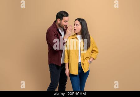 Young man whispering secret to shocked girlfriend looking at him with mouth open while standing isolated on beige background. Couple sharing mysteriou Stock Photo