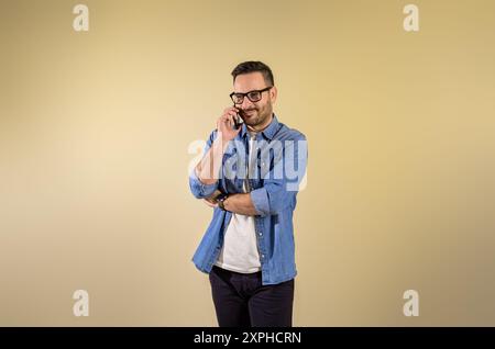 Smiling confident businessman talking over mobile phone and standing isolated against beige background. Happy young man wearing denim shirt and eyegla Stock Photo