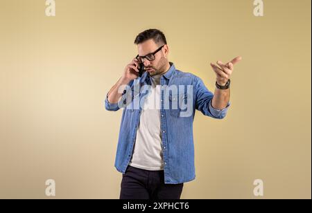 Frustrated male entrepreneur discussing serious problem over mobile phone. Stressed young man making call and gesturing against isolated beige backgro Stock Photo