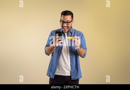 Male customer with credit card and smart phone screaming excitedly after winning rewards for online shopping. Cheerful young man buying through mobile Stock Photo