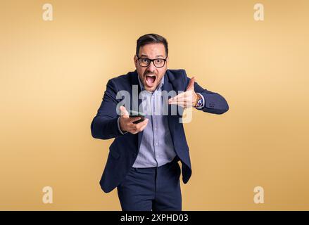 Portrait of angry stressed male manager pointing at mobile phone and shouting furiously against yellow background. Frustrated businessman holding cell Stock Photo