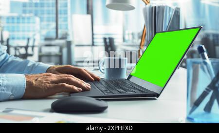 Close-Up On Hands Of Professional Business Manager Using Laptop Computer With Green Screen Chromakey On Display. Man Analyzing Financial Reports And Enjoying Hot Coffee In The Corporate Office. Stock Photo
