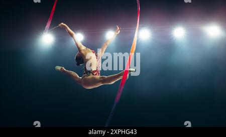 Graceful Female Gymnast in Mid-air, Performing Split Leap With Ribbon Under Bright Stadium Lights. Flexibility, Elegance, and Precision in Captivating Rhythmic Gymnastics Routine. Stock Photo