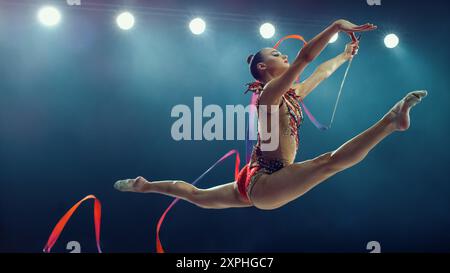 Asian Female Gymnast in Mid-air, Performing Dynamic Split Leap With Ribbon Under Bright Stadium Lights. Elegance, Flexibility, and Precision in an Exhilarating Rhythmic Gymnastics Routine. Stock Photo