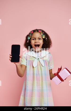 A young African American girl in a colorful dress smiles and holds a phone with a blank screen. Stock Photo