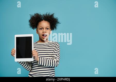 A young African American girl with curly hair holds up a tablet with a surprised expression on her face. Stock Photo