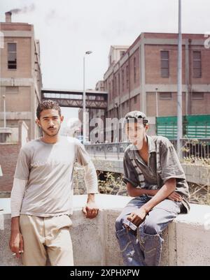 Two young male textile factory workers taking a break in Alexandria, Egypt. The image captures an industrial setting with factory buildings in the background. June 2006. Stock Photo