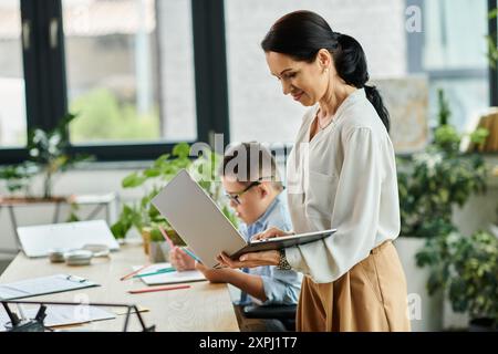 A mother works on her laptop while her son with Down syndrome works on his schoolwork at their home office. Stock Photo