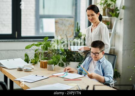 A mother works on her laptop while her son, who has Down syndrome, draws at the table in a home office setting. Stock Photo