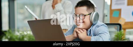 A young boy with Down syndrome wearing headphones works on a laptop while his mother works in the office next to him. Stock Photo