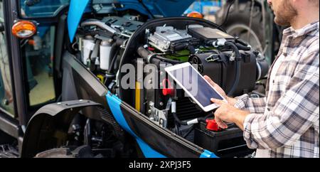 Mechanic diagnoses tractor engine operation using digital tablet. Blank screen of digital tablet in hands in front of machinery engine. Stock Photo