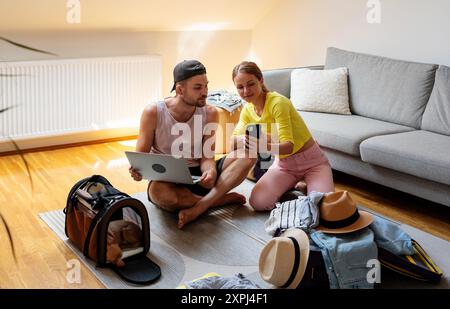 Young married couple sit on the floor in room using phone and laptop for booking travel. Stock Photo