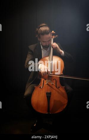 Vertical full shot of male musician playing cello with concentration looking down on string instrument while sitting in dark hazy studio with dim top light Stock Photo