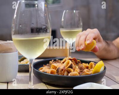 A hand squeezing a lemon over a plate of fried squids with two glasses of white wine Stock Photo