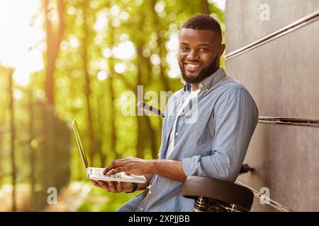 Black smiling freelancer holding laptop outside and leaning on bicycle Stock Photo