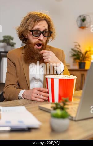 Caucasian businessman freelancer taking break from work wearing 3D glasses earing popcorn and watching movie on laptop. Smiling man guy sitting at home office desk having snacks during break. Vertical Stock Photo
