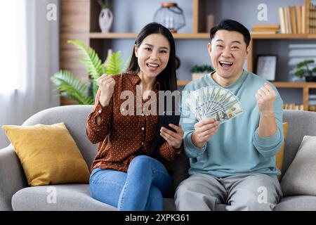 Asian couple celebrating financial success while sitting on couch holding smartphones and cash. They are happy and excited, showing positive expressions and gestures Stock Photo