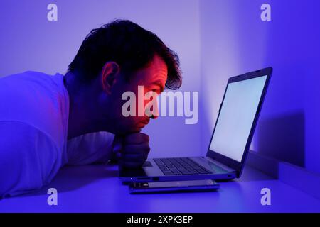A man sits closely in front of a laptop, deeply concentrated on the bright screen. His brow is furrowed, and he is supporting his chin with his hand, Stock Photo