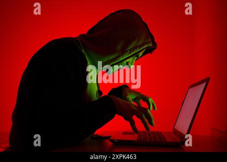 A person dressed in a hooded sweatshirt is intently focused on a laptop in a dimly lit setting. The individual s silhouette is illuminated in green, c Stock Photo
