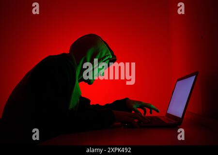 An individual dressed in a green hoodie is intently focused on a laptop, sitting in a dimly lit environment with a striking red background. The person Stock Photo