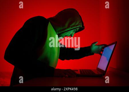 An individual wearing a green and black hoodie sits in front of a laptop, visibly frustrated. The room is illuminated with a striking red light that c Stock Photo