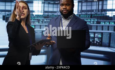Teamworking colleagues doing checkup, inputting data on laptop of needed server hub fixes. Coworkers working together in data center housing advanced storage infrastructure, camera A Stock Photo