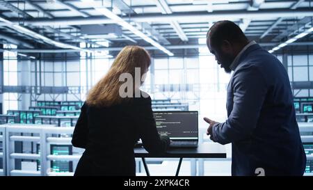 Team of developers doing maintenance checkup using laptop to prolong data center hardware life span. IT support staff members in server hub monitor server infrastructure using notebook, camera B Stock Photo