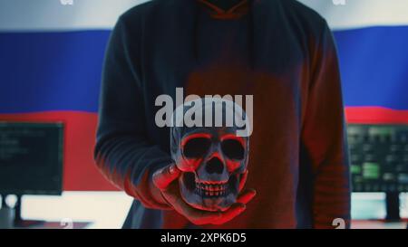A maniacal russian hacker creating cyber threat alert by holding a skull as concept of cyber espionage and malicious code. Illustration warning of cyber security risks, evil harmful scam. Camera B. Stock Photo
