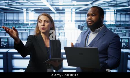Teamworking colleagues doing checkup, inputting data on laptop of needed server hub fixes. Coworkers working together in data center housing advanced storage infrastructure, camera B Stock Photo