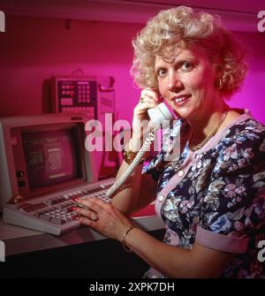 Retro office scene with female operator using landline phone and early computer terminal, bathed in neon pink lighting, evoking 1980s tech era. Stock Photo