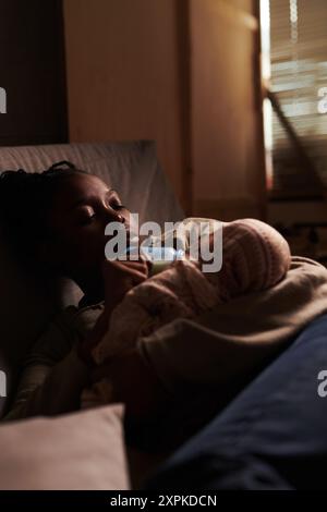 Young tired woman with baby on hands lying on couch in front of camera in home environment and feeding infant with milk Stock Photo