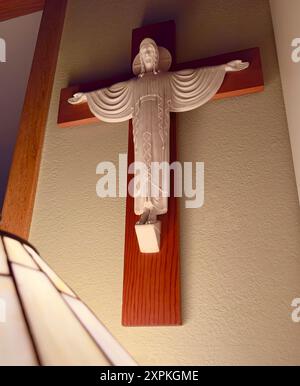 Cross crucifix sculpture hangs on plaster wall with wood framework. Light from lamp shade shines upward on face of Jesus. Jesus is light of the world. Stock Photo