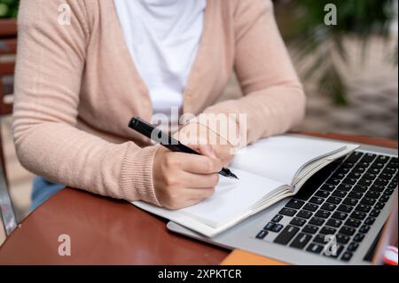 A cropped image of a woman in casual clothes making a list or taking notes in her notebook while studying online on her laptop at an outdoor table. Stock Photo