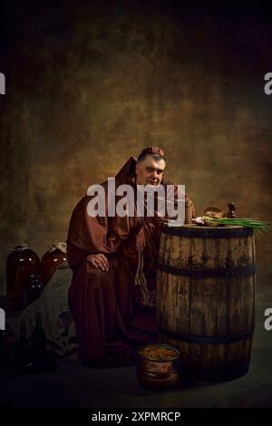 Portrait of medieval man, monk in period attire leaning on wooden barrel with wooden mug of beer against vintage dark background. Stock Photo