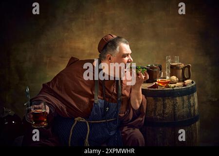 Man, monk in period attire enjoying sandwich with fish and drinking ale, against vintage background with wooden barrel with various beer mugs on top Stock Photo