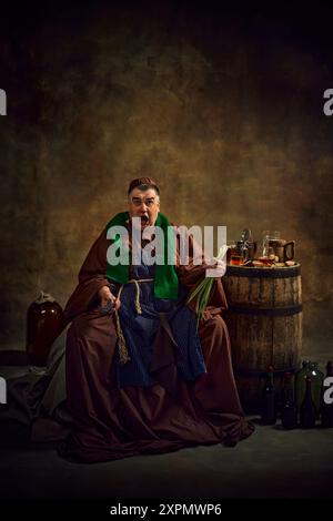 Sport fan supporting soccer team. Emotional monk in period attire watching game, drinking beer gain dimly lit vintage background. Stock Photo